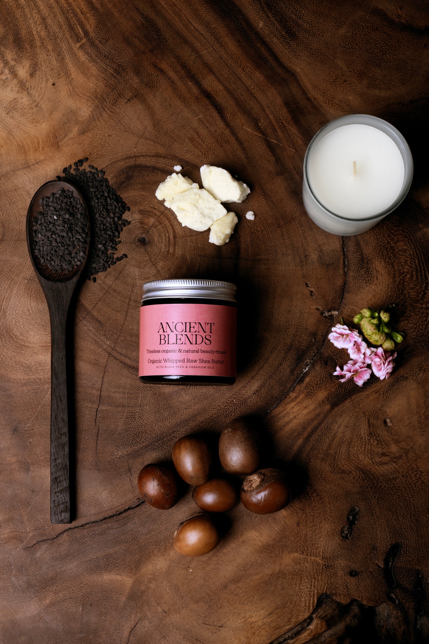 Candle, wooden spoon, shea butter, geranium flowers and black seed on a wooden surface with 'Ancient Blends' product.