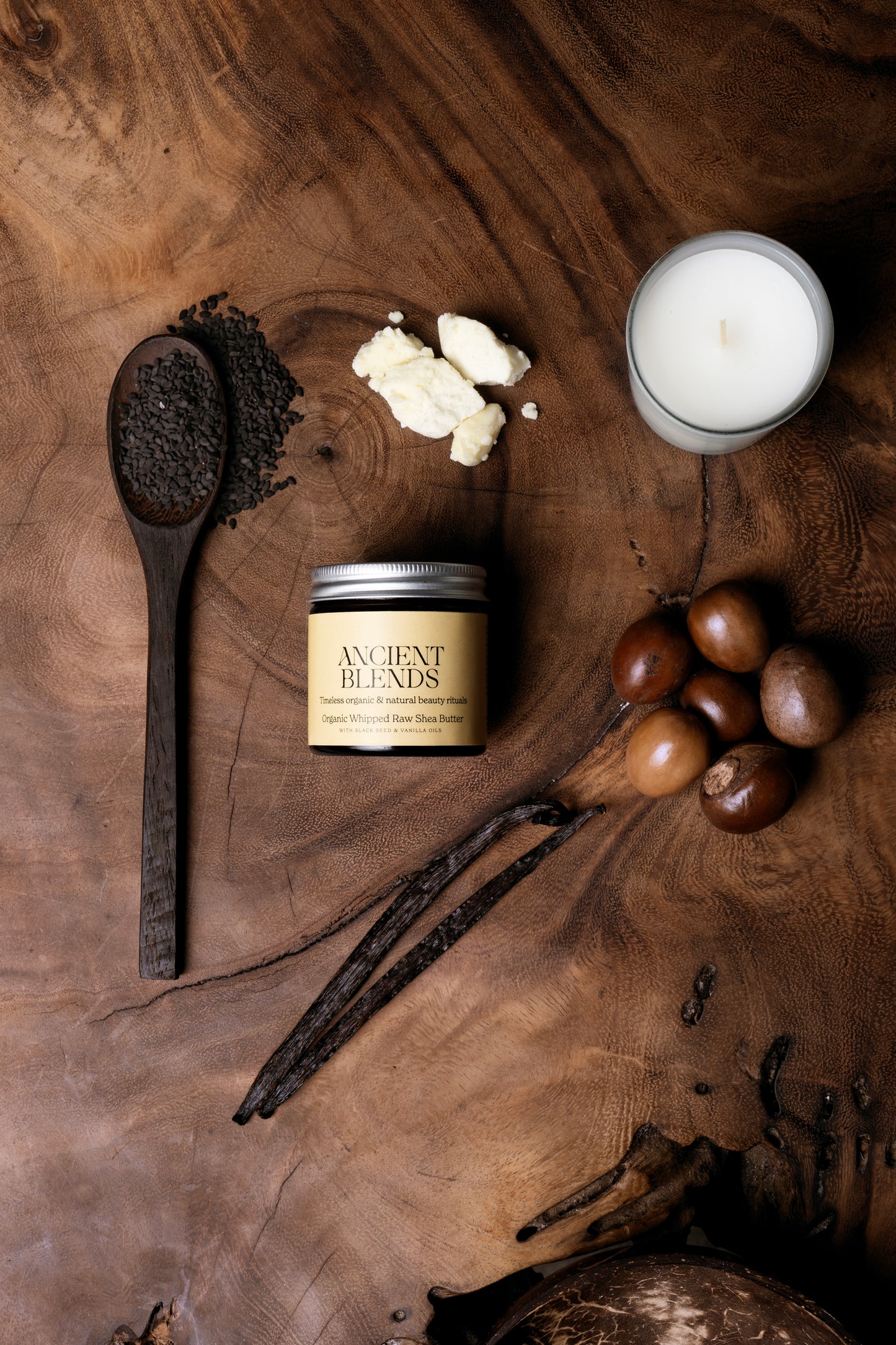 Candle, wooden spoon, shea butter, vanilla beans and black seed on a wooden surface with 'Ancient Blends' product.